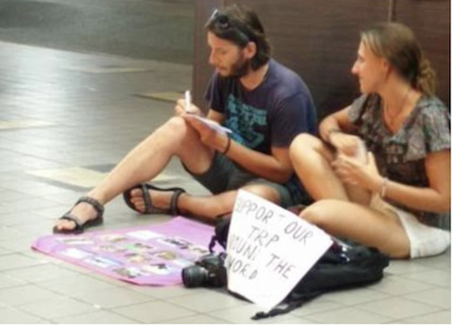 صورة لشاب وصديقته في سنغافورة يجلسان على الأرض في مكان عام ويحملان لافتة كتب عليها ادعموا رحلتنا حول العالم